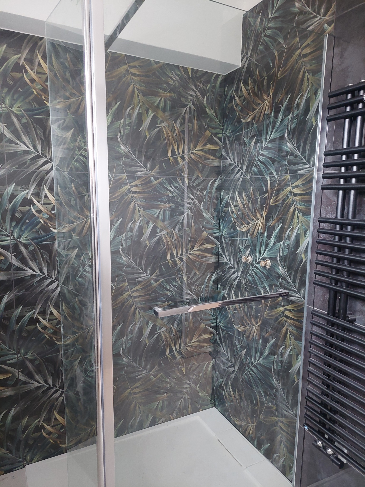 Nowoczesna kabina prysznicowa z deszczownicą, wykończona płytkami w tropikalne liście. Czarny grzejnik drabinkowy po prawej stronie. Szklane drzwi kabiny.
