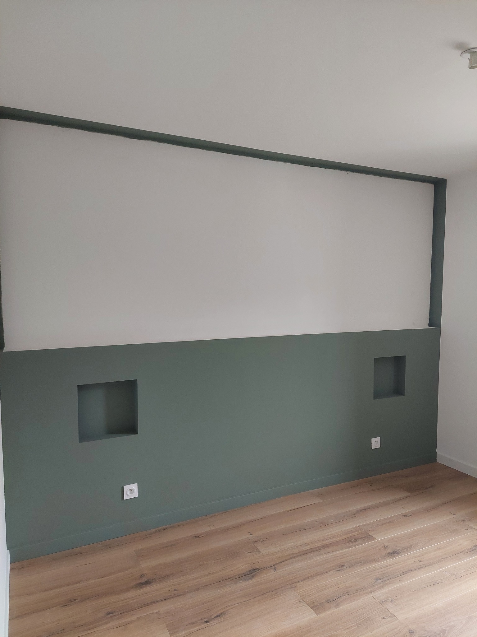 Dwukolorowa ściana z wnękami i listwami w odcieniach zieleni, uzupełniona drewnopodobną podłogą i gniazdkami elektrycznymi. Minimalistyczny design wnętrza.