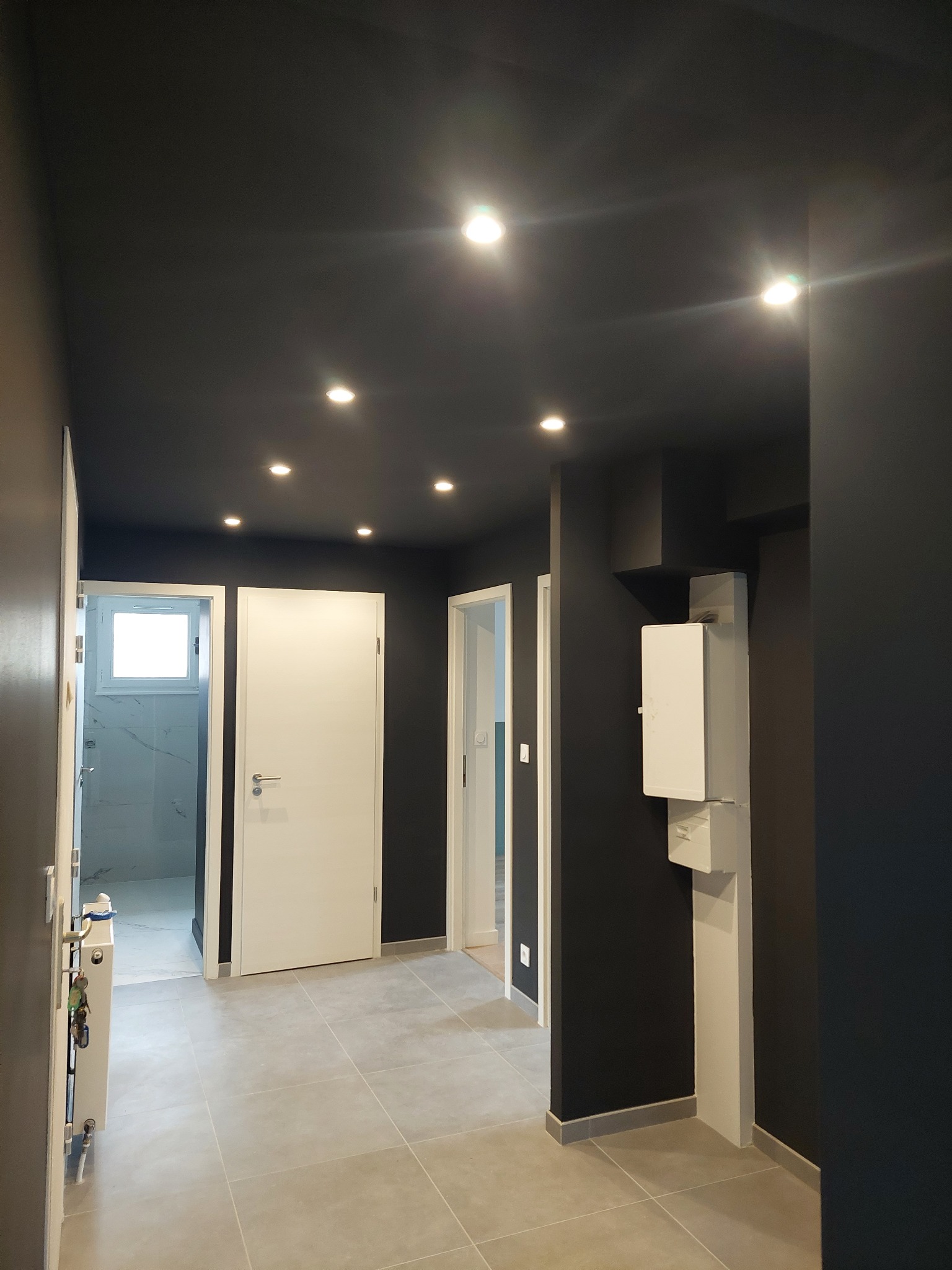 Nowoczesny korytarz po remoncie: ciemne ściany i sufit, jasna podłoga z płytek, białe drzwi i oświetlenie punktowe. Widok na łazienkę z marmurowymi płytkami.