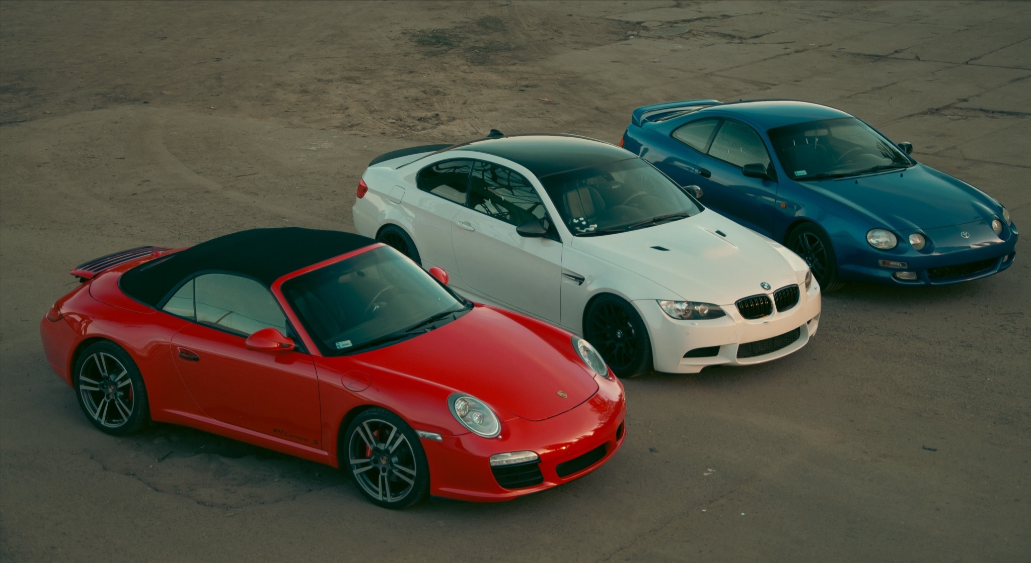 Trzy sportowe auta: czerwone Porsche 911 Cabrio, białe BMW M3 z czarnym dachem i niebieska Toyota Celica, stojące na betonowym placu, sfotografowane z góry.
