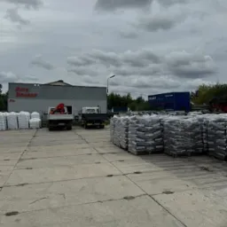 Skład opału z zapasem workowanego węgla i ekogroszku na placu, ciężarówki w tle. Widok na budynek firmy Auto Dragon.