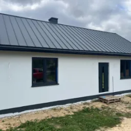 Nowoczesny, biały dom z szarą, metalową blachodachówką i ciemnymi oknami. Widoczna paleta pod drzwiami. Tło: pochmurne niebo.