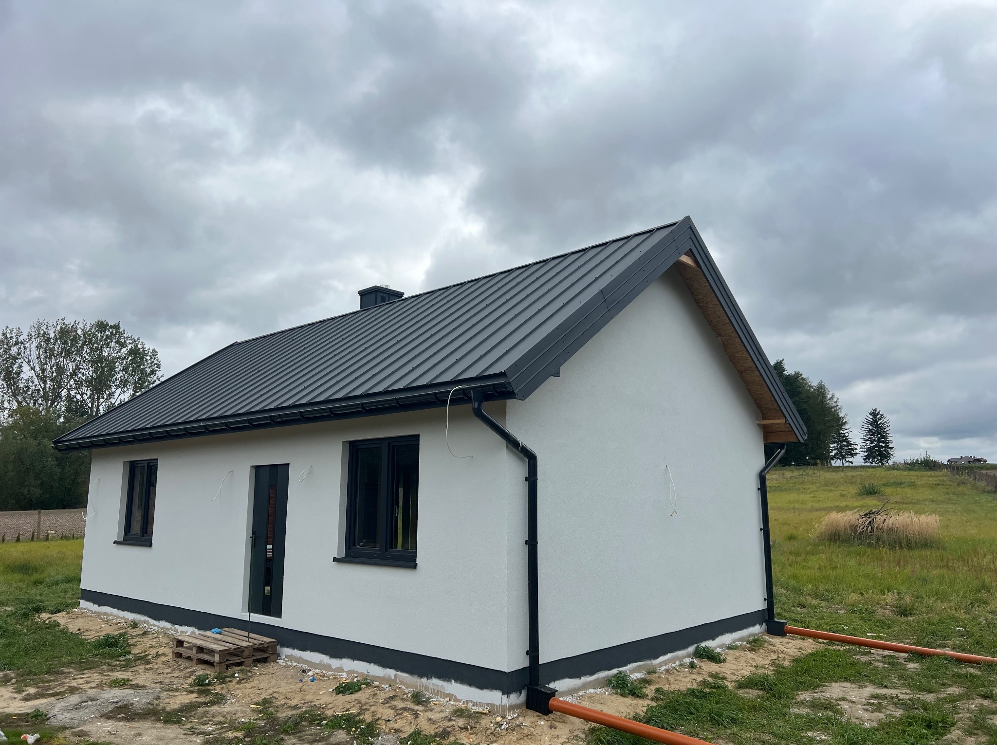 Niewielki, parterowy dom z szarą blachą na dachu i białymi ścianami, z widocznymi rynnami i rurami spustowymi. Na ziemi leżą palety.