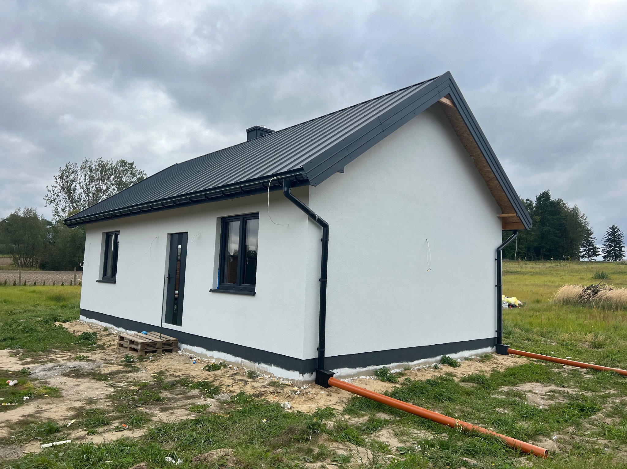Gotowy, parterowy dom z szarą blachodachówką, białymi ścianami i czarnymi oknami. Widoczne rury odprowadzające wodę i palety przy wejściu. Pochmurne niebo w tle.