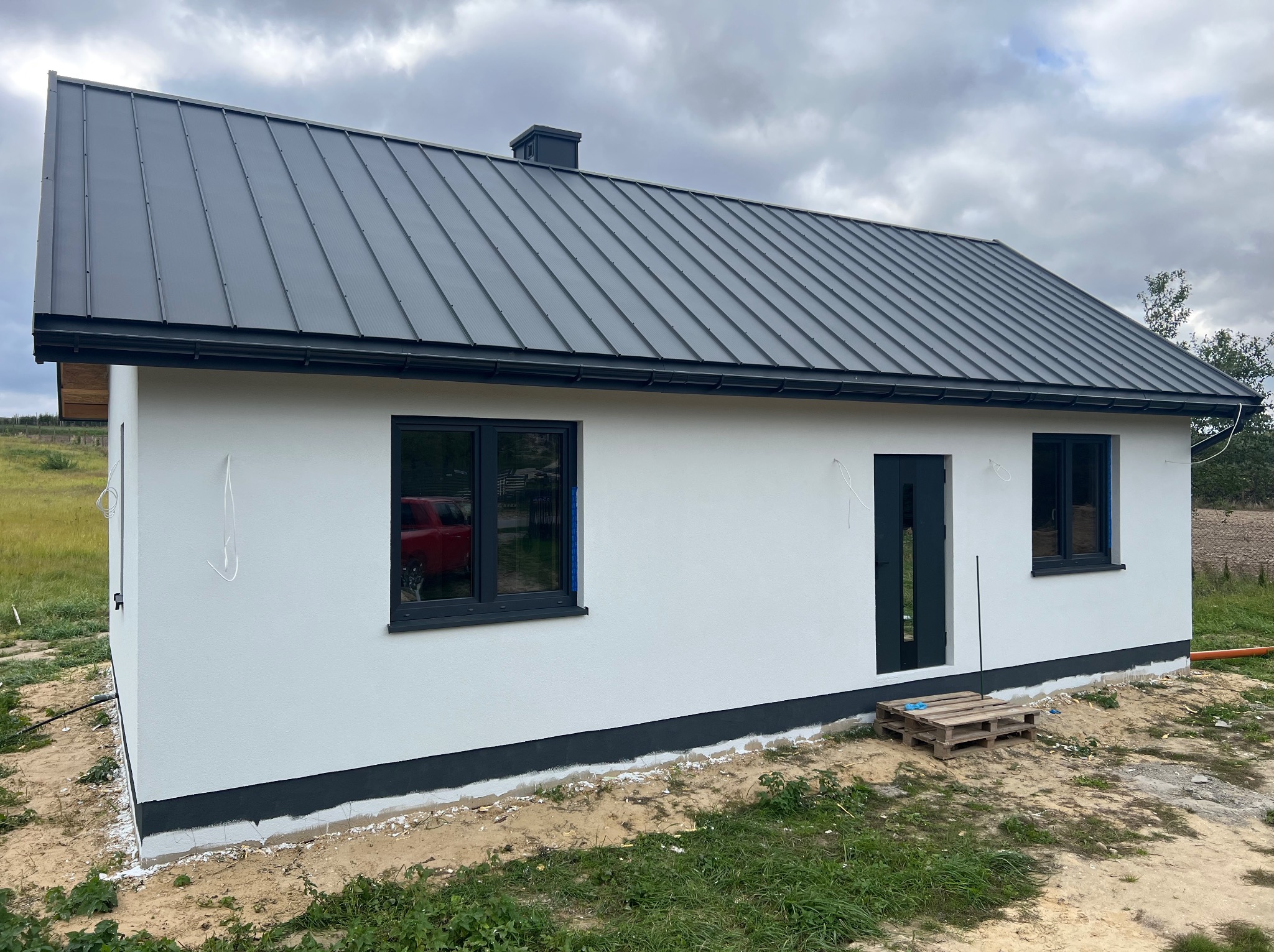 Nowoczesny, biały dom z szarą, metalową blachodachówką i ciemnymi oknami. Widoczna paleta pod drzwiami. Tło: pochmurne niebo.