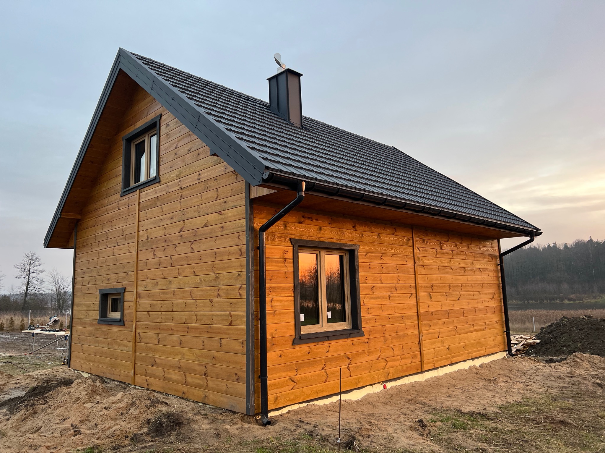 Złocisty dom z drewnianą elewacją i ciemnym dachem z blachy, widoczny z boku na tle zachodzącego słońca. Okna z ciemnymi ramami odbijają ciepłe światło.