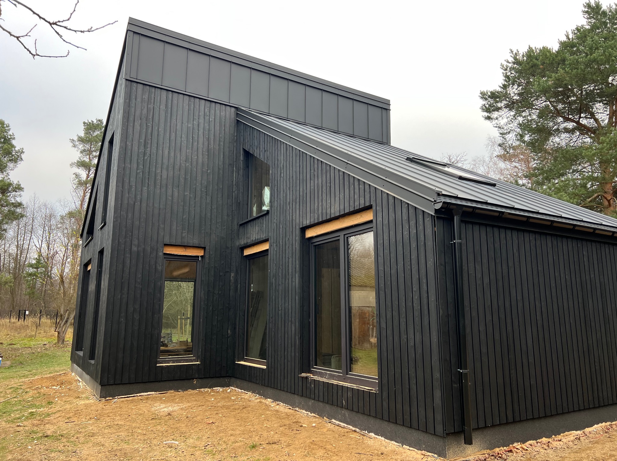 Nowoczesny, czarny dom z drewnianą elewacją i metalowym dachem. Widoczne okna z drewnianymi ramami i rynna. Dom otoczony ziemią i drzewami.