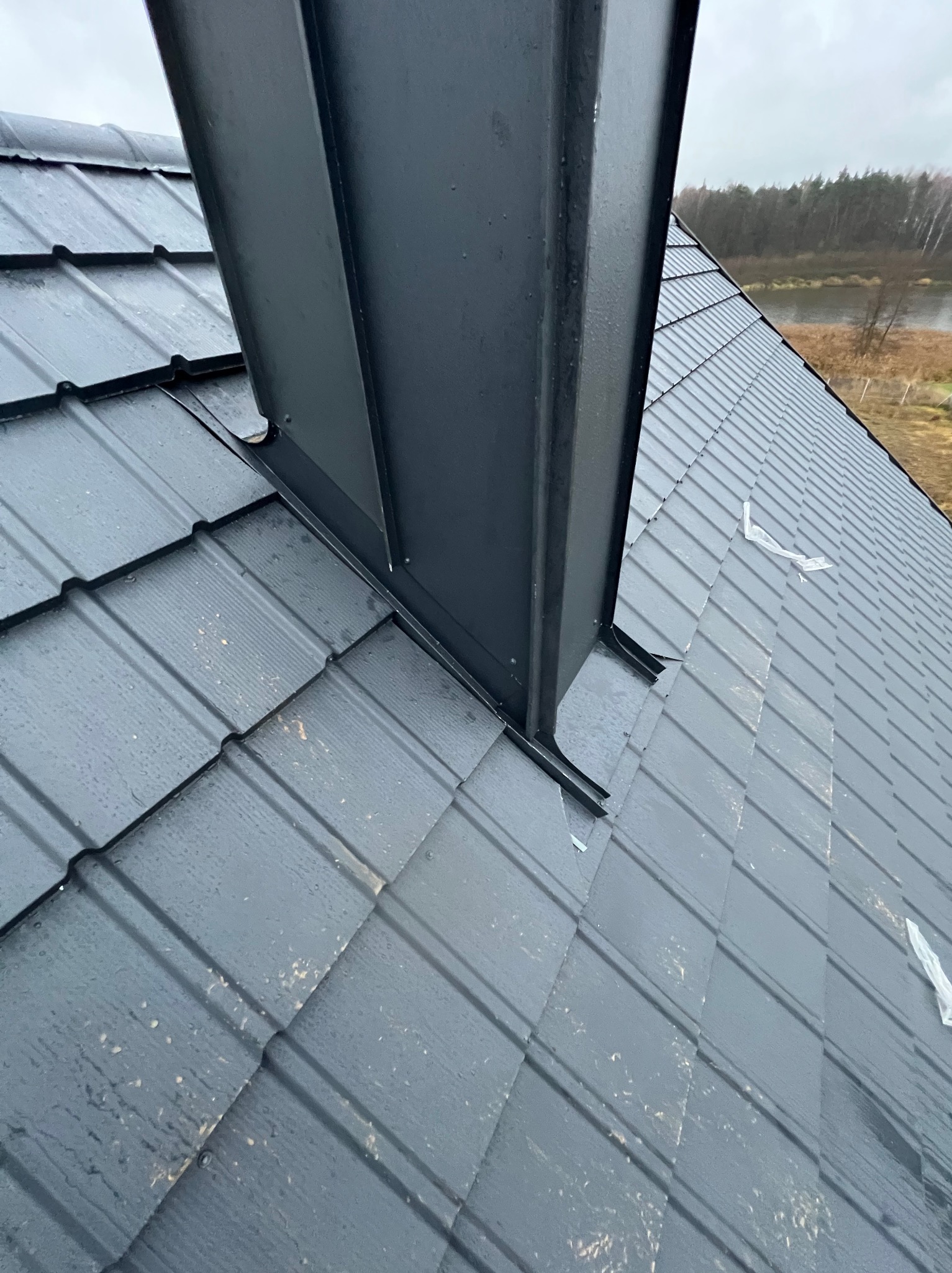 Szczegółowe ujęcie czarnego dachu z blachy trapezowej z kominem, widoczne ślady po deszczu, w tle zalesiony krajobraz. Kątowe ujęcie podkreśla strukturę pokrycia.