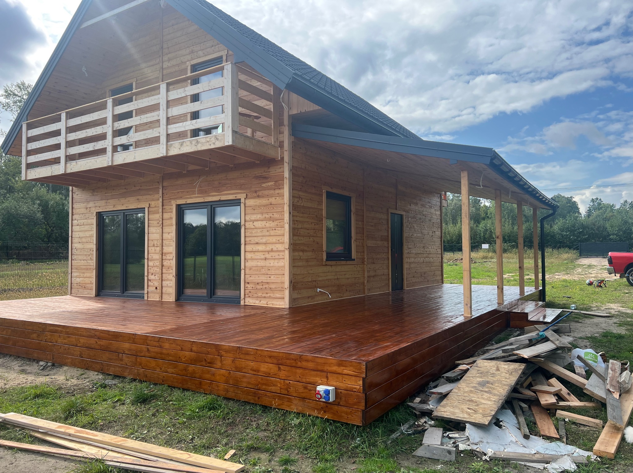 Nowoczesny dom szkieletowy z drewnianym tarasem i balkonem. Widoczne czarne okna i grafitowy dach. Na pierwszym planie deski i odpady budowlane.