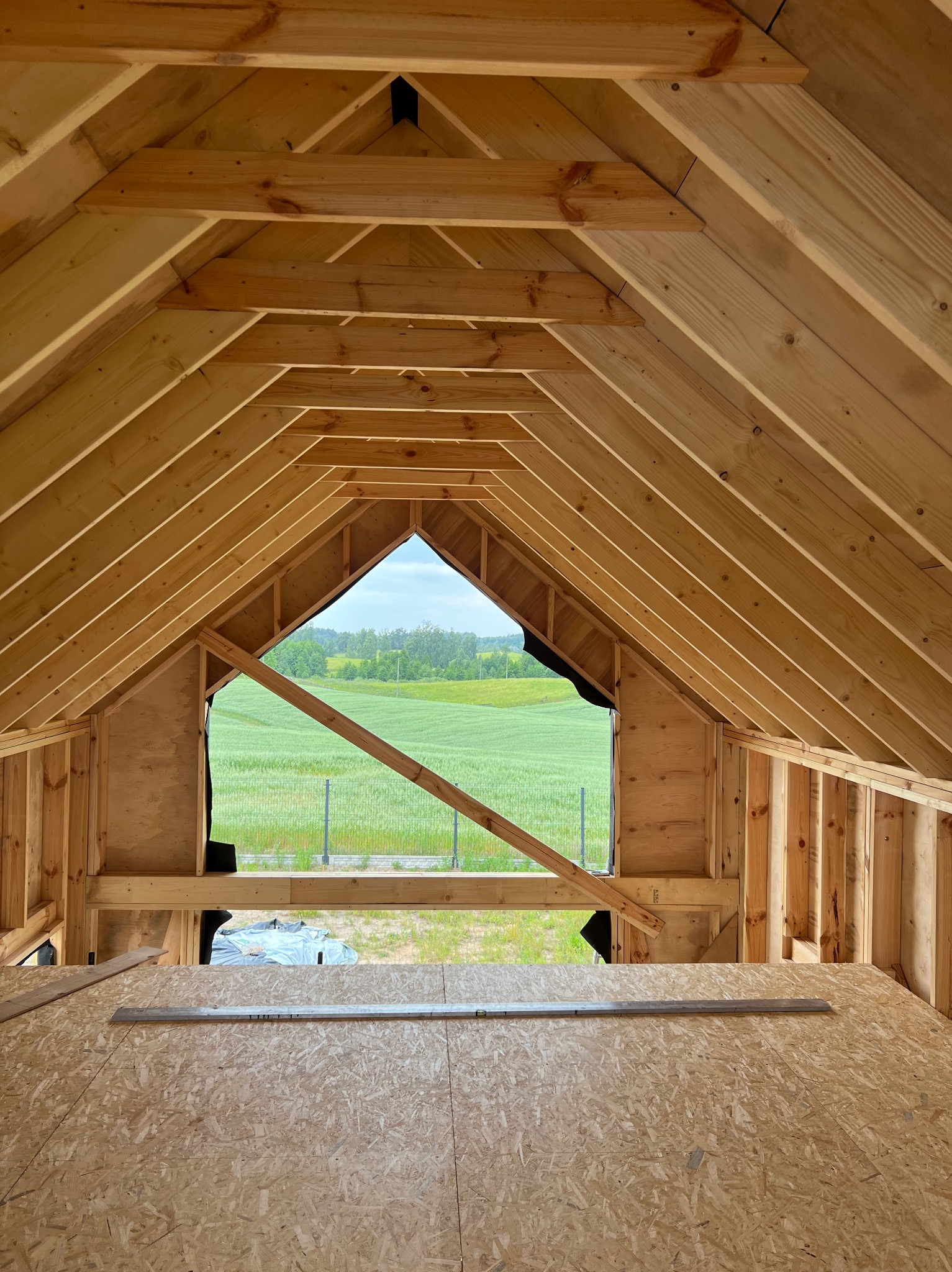 Wnętrze domu w trakcie budowy, widok na drewnianą konstrukcję dachu i podłogi z płyty OSB, z oknem na zielone pole w tle. Widoczny etap budowy.