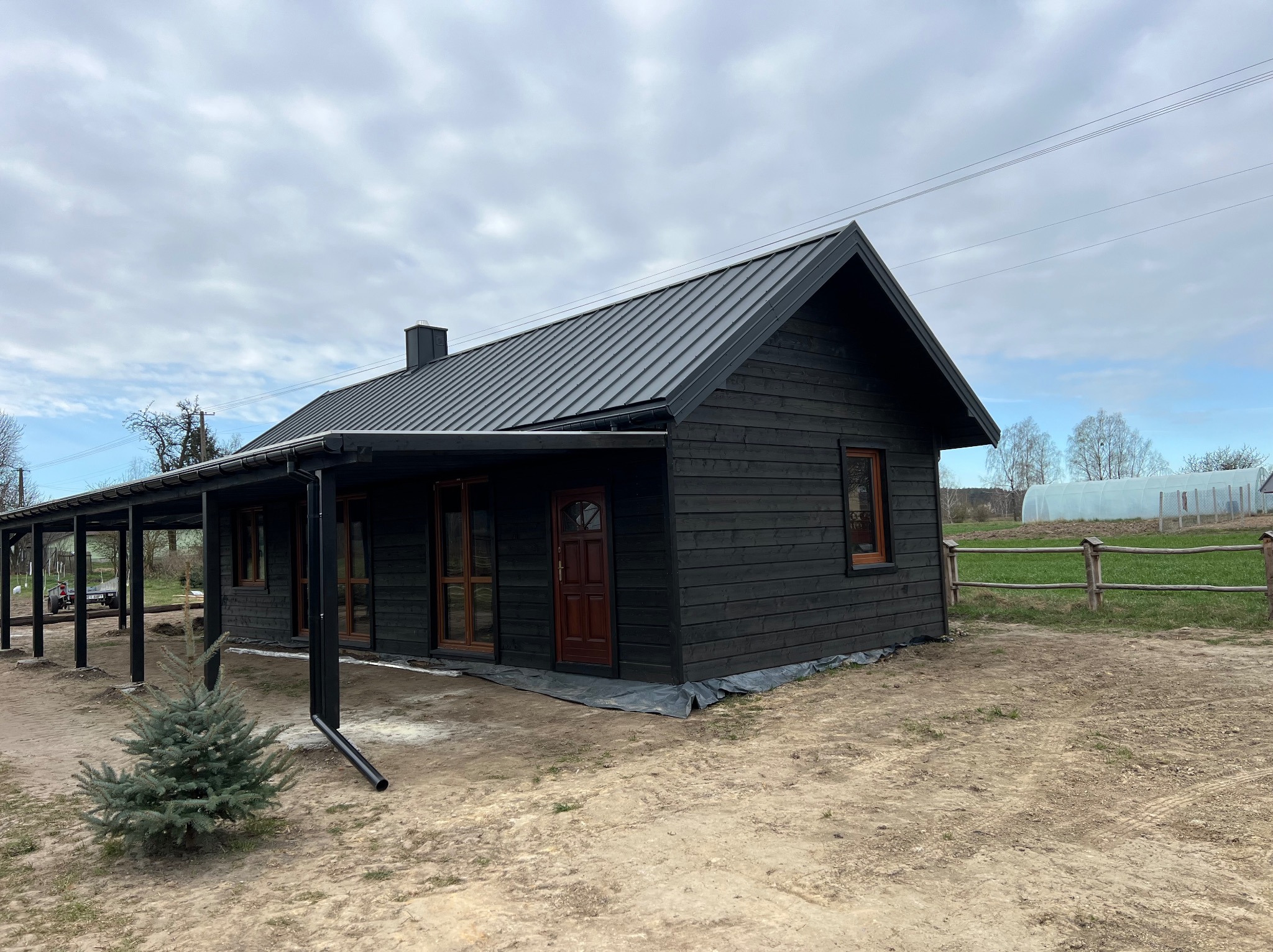 Czarny dom z drewnianą stolarką okienną i metalowym dachem na działce. Widoczny fragment zadaszonego tarasu i mała choinka. W tle szklarnia i drewniane ogrodzenie.