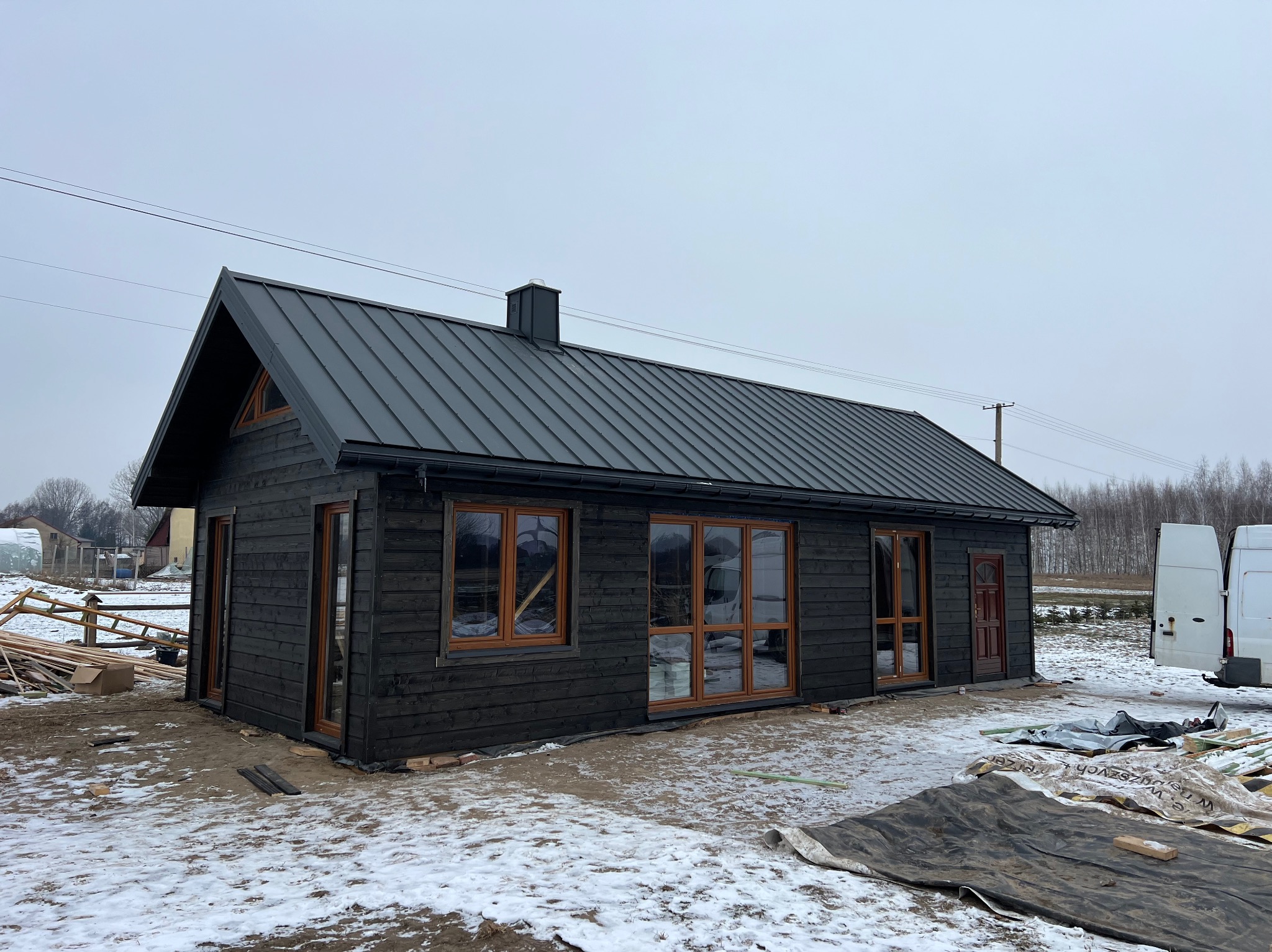 Czarny dom z drewnianą elewacją i metalowym dachem w zimowej scenerii. Widoczne okna z drewnianymi ramami i elementy wykończeniowe. Częściowo ośnieżony teren.