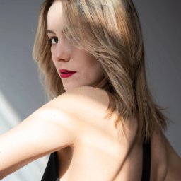 Karolina Sabik Photography - Portret kobiety z blond włosami, odwróconej przez ramię, z czerwonymi ustami i delikatnym makijażem, w naturalnym świetle na szarym tle.