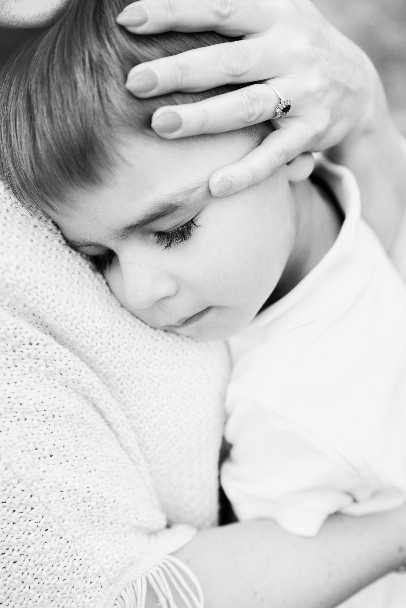 Czarno-białe zdjęcie z bliska: chłopiec wtulony w matkę, jej dłoń z pierścionkiem delikatnie spoczywa na jego głowie. Spokój i bliskość w intymnym ujęciu.