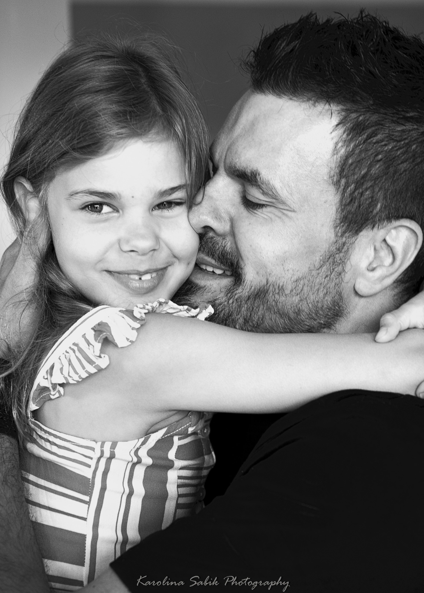 Czarno-białe ujęcie uśmiechniętej dziewczynki wtulonej w tatę, który czule ją obejmuje. Ciepła relacja ojca i córki uchwycona w bliskim kadrze, z delikatnym oświetleniem.