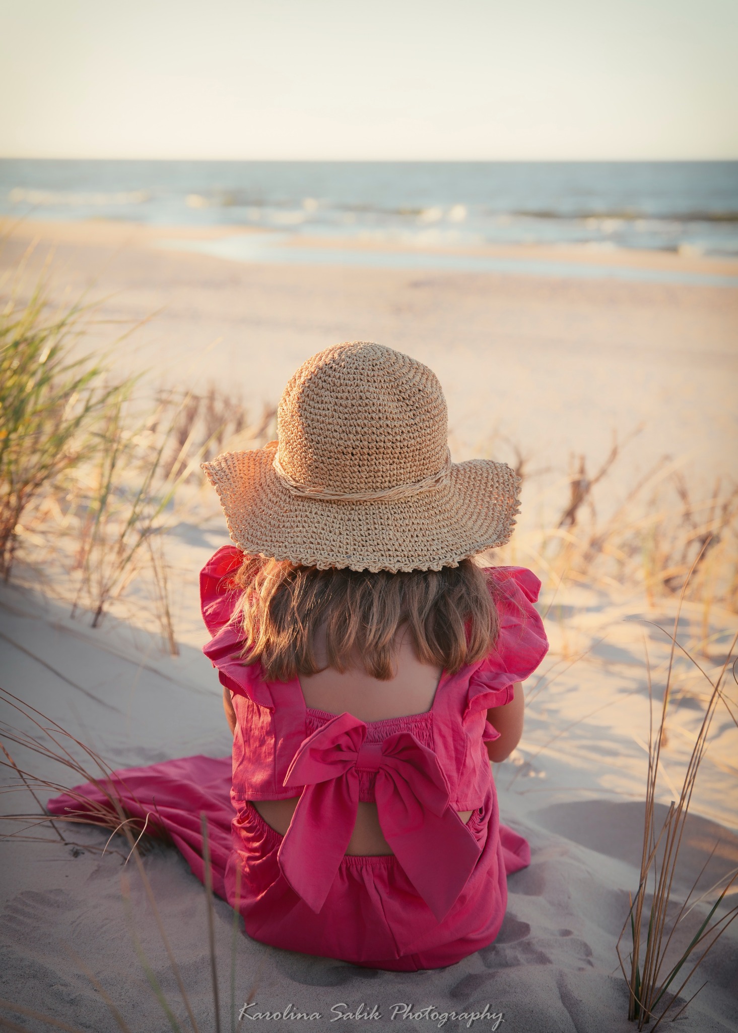 Dziewczynka w różowej sukience i słomkowym kapeluszu siedzi na piaszczystej plaży, tyłem do widza, patrząc na morze w ciepłym, zachodzącym słońcu.