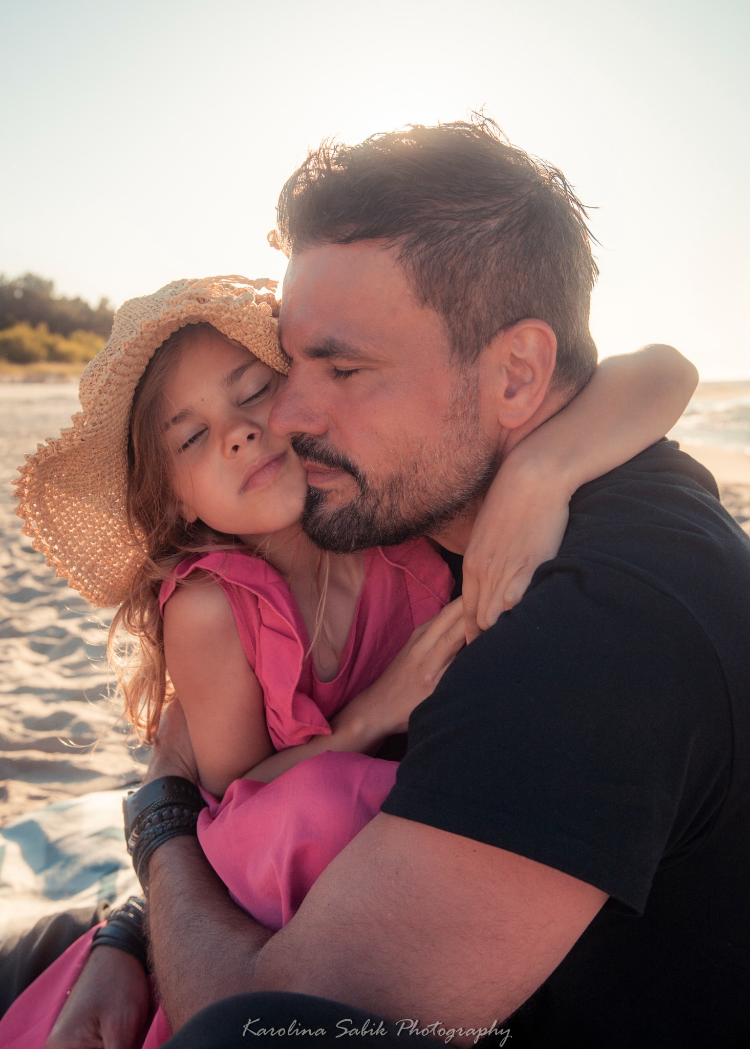 Ojciec tuli córkę na plaży o zachodzie słońca. Dziewczynka w słomkowym kapeluszu i różowej sukience wtula się w tatę. Ciepłe, naturalne światło, spokojna atmosfera.
