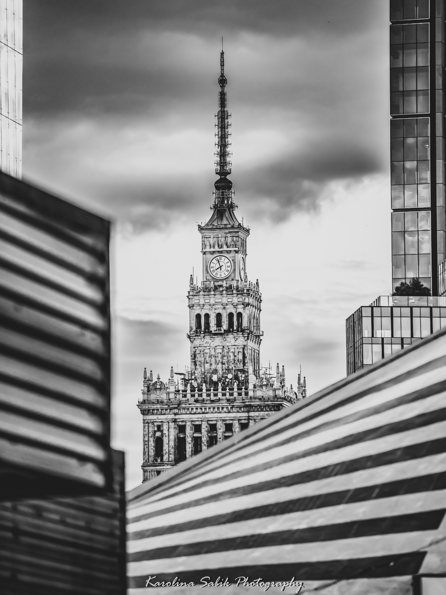 Pałac Kultury w Warszawie, uchwycony w czerni i bieli, z perspektywy między elementami konstrukcyjnymi. W tle nowoczesne budynki i pochmurne niebo.