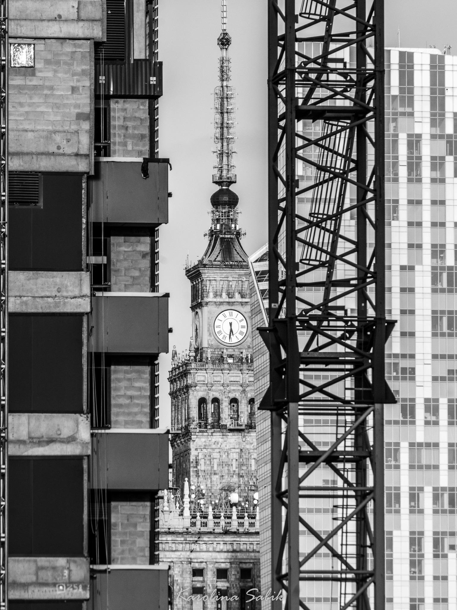 Pałac Kultury i Nauki w Warszawie widziany pomiędzy konstrukcją budynku w budowie i żurawiem, czarno-biała fotografia z perspektywy ulicy.