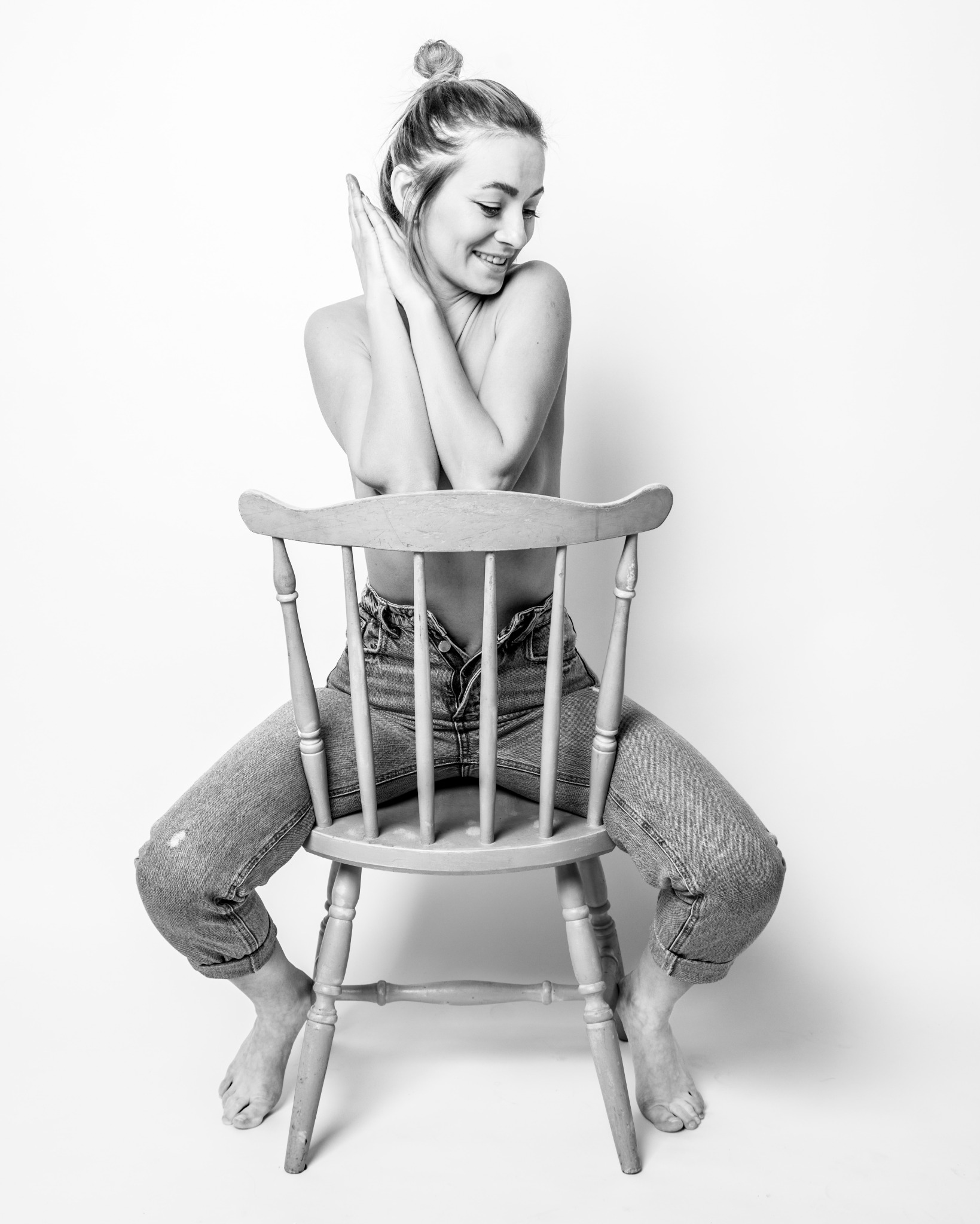 Czarno-białe zdjęcie kobiety siedzącej na krześle, ubrana w dżinsy, z rękami złożonymi przed sobą, spoglądającej w dół z uśmiechem, na jasnym tle.