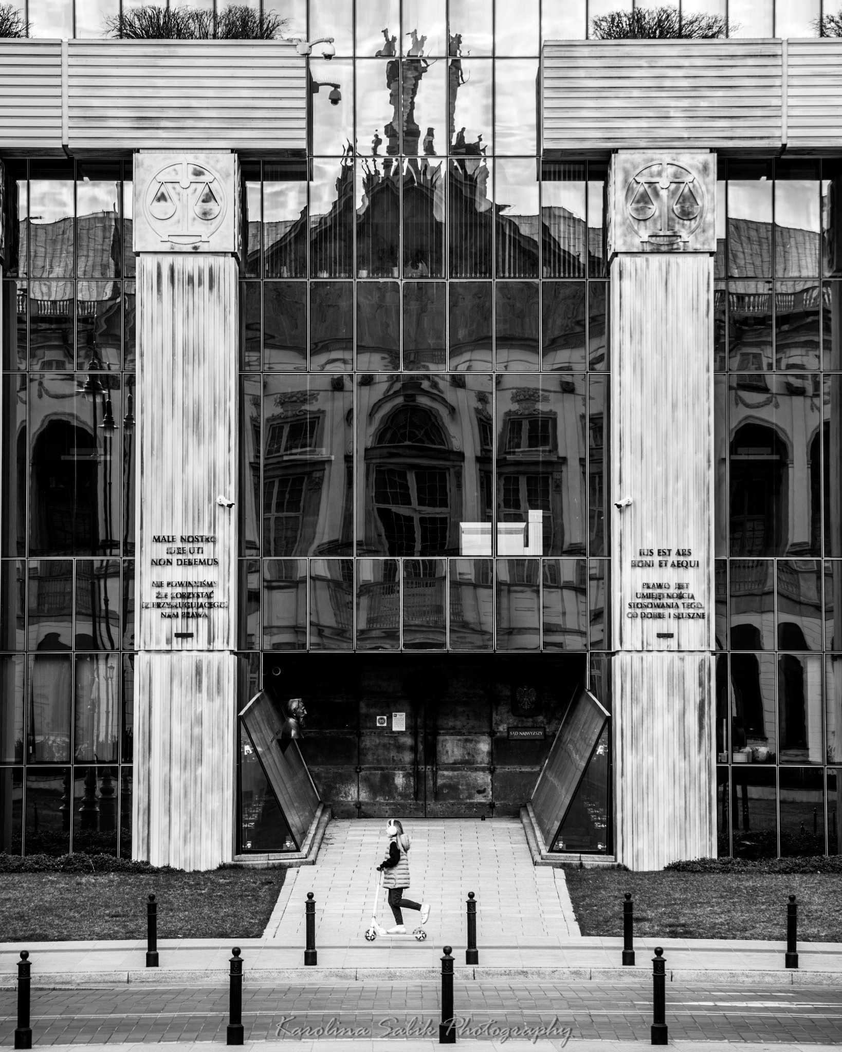 Czarno-białe zdjęcie Sądu Najwyższego w Warszawie z odbiciem budynku w szklanej fasadzie i dziewczynką na hulajnodze przed wejściem. Architektura modernistyczna.