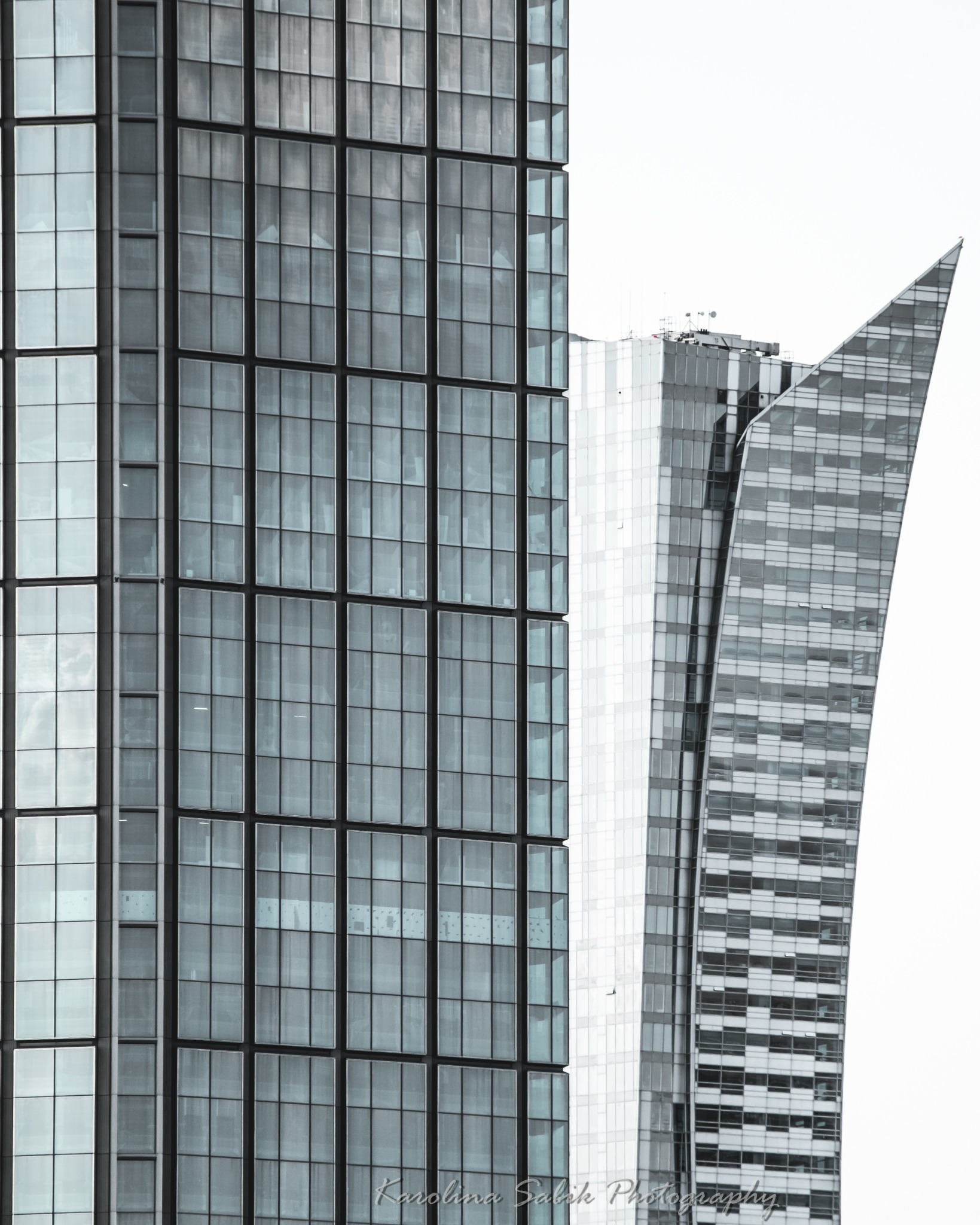 Abstrakcyjne ujęcie trzech wieżowców o zróżnicowanej architekturze, z dominującą szklaną fasadą w odcieniach szarości i bieli, na tle jasnego nieba. Minimalizm i nowoczesność.