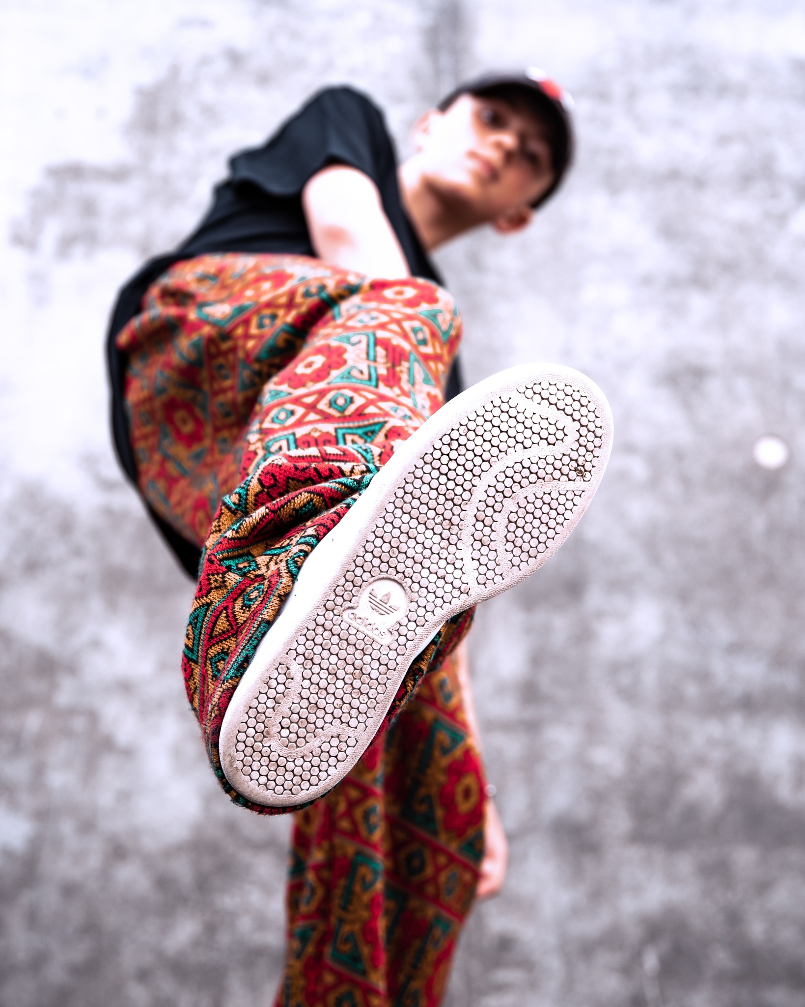 Osoba w czarnej koszulce i wzorzystych spodniach prezentuje podeszwę buta Adidas Stan Smith na tle szarej ściany. Nietypowy kąt ujęcia.
