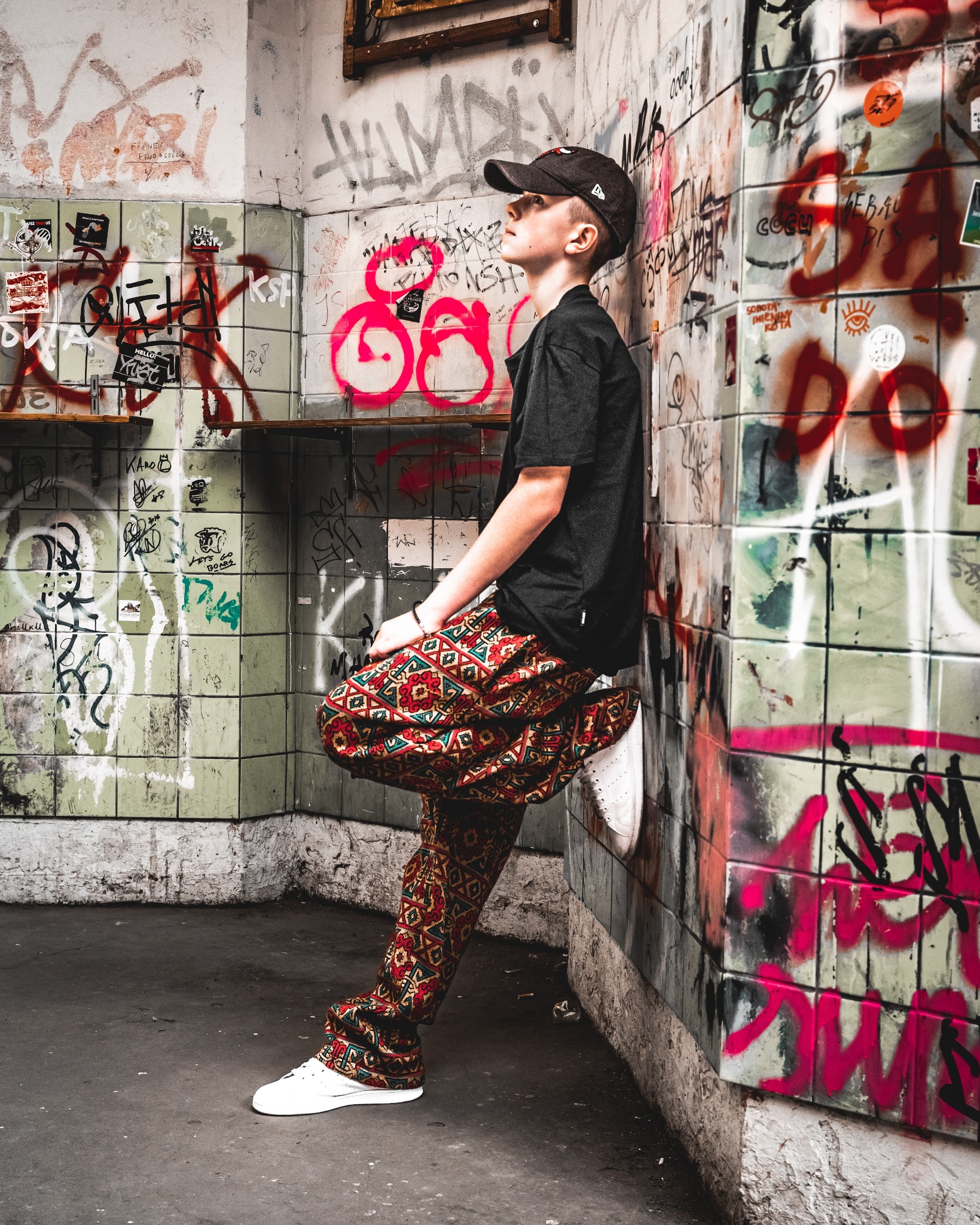 Młody mężczyzna w czarnej koszulce i wzorzystych spodniach opiera się o ścianę pokrytą graffiti w opuszczonym wnętrzu. Patrzy w górę. Ujęcie z dołu.