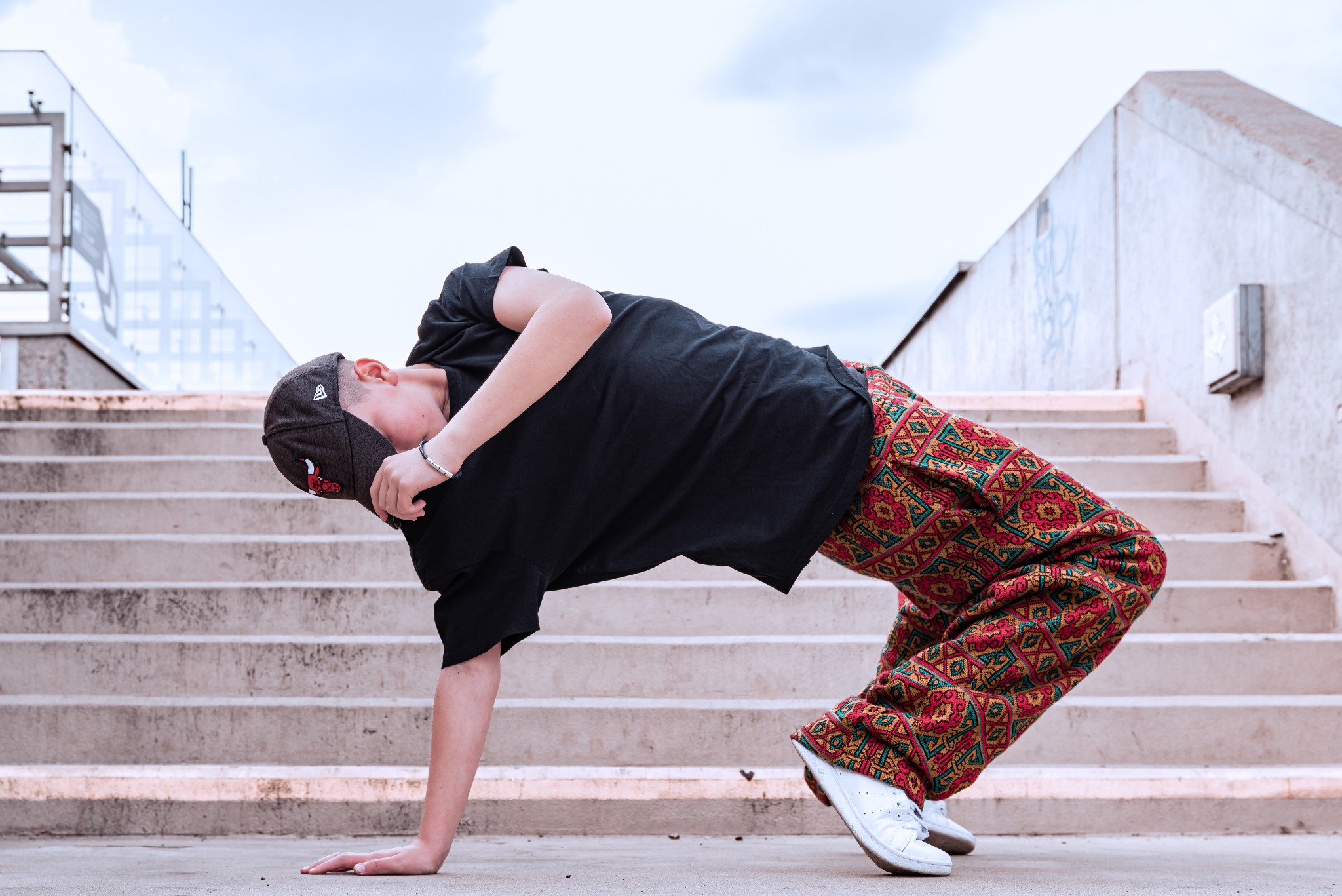 Młody mężczyzna w oryginalnych spodniach i czarnej koszulce wykonuje skomplikowaną figurę taneczną na tle betonowych schodów i nieba.