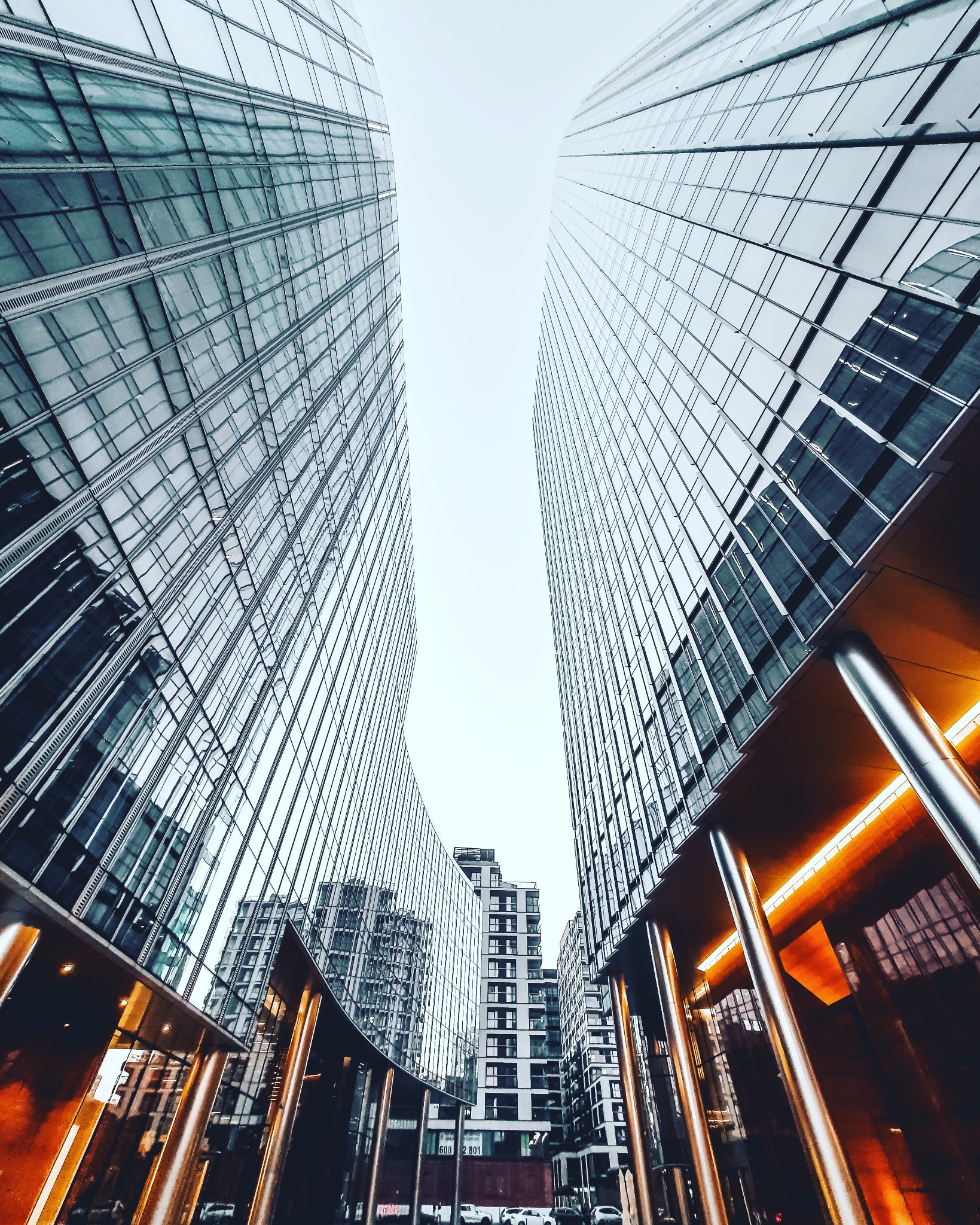 Wertykalny kadr na dwa nowoczesne, szklane biurowce z widocznymi odbiciami innych budynków w fasadach. Ujęcie z dołu, podkreślające wysokość i architekturę.