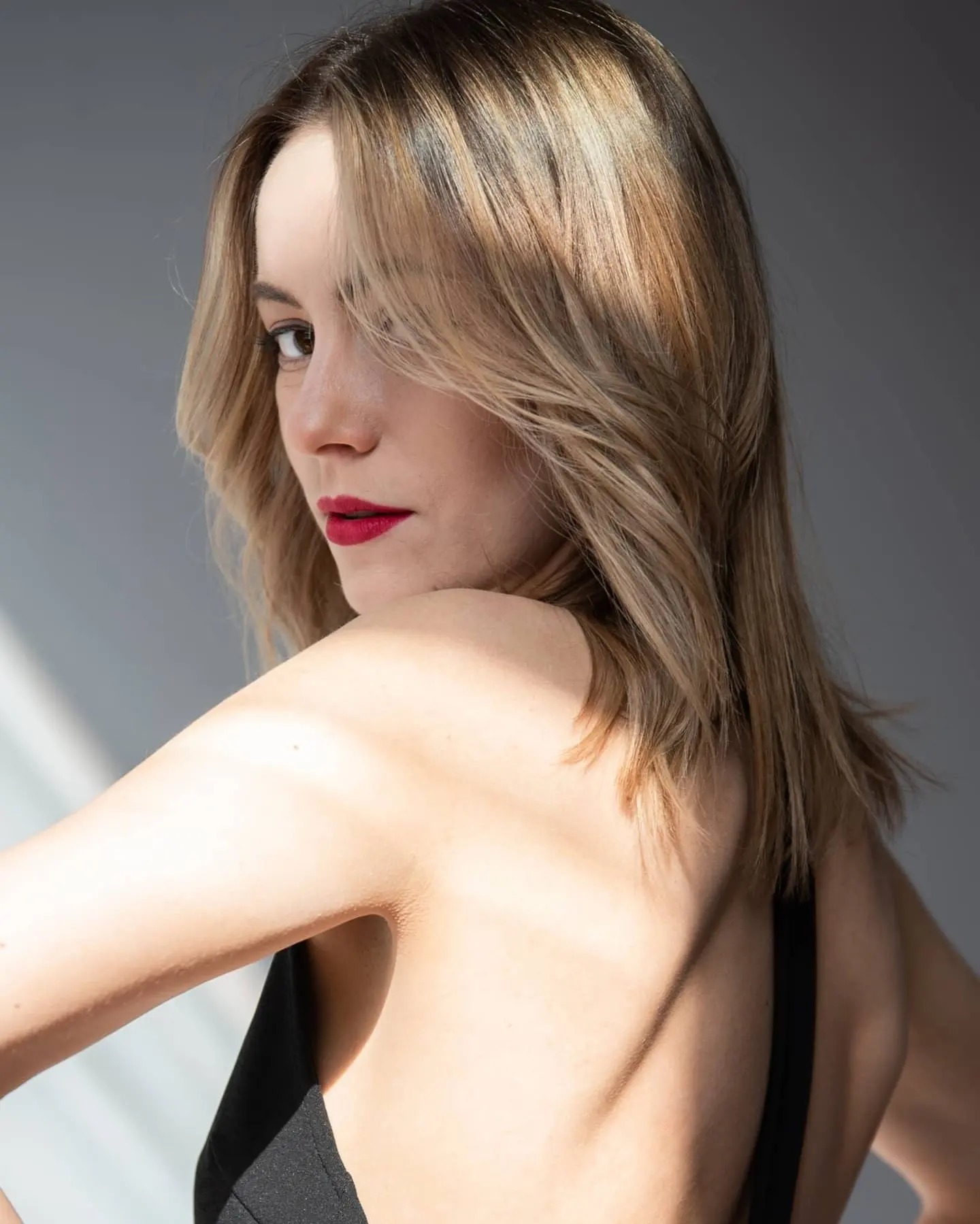 Portret kobiety z blond włosami, odwróconej przez ramię, z czerwonymi ustami i delikatnym makijażem, w naturalnym świetle na szarym tle.