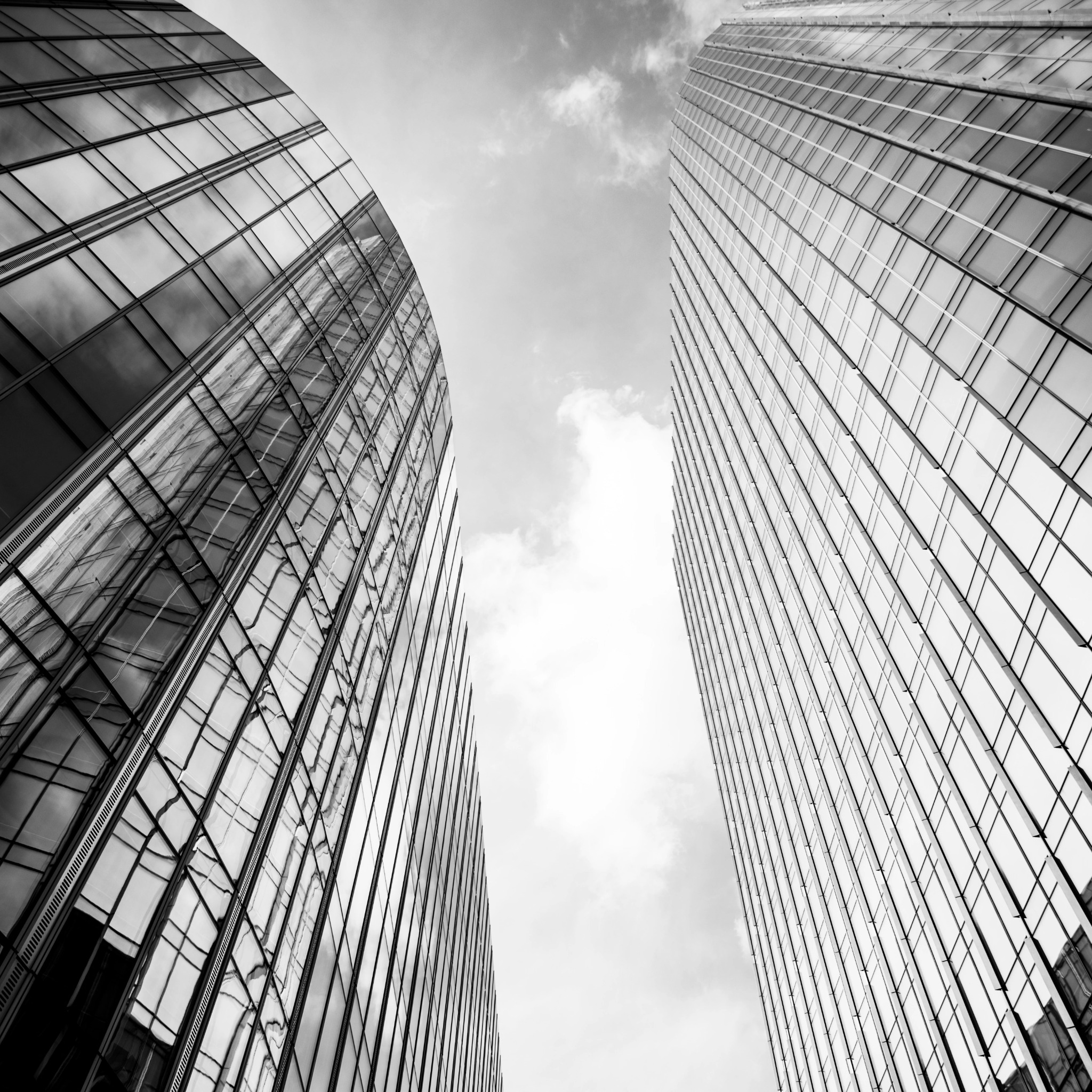 Monochromatyczne ujęcie z dołu na dwie nowoczesne, szklane wieże biurowe, z refleksami nieba i chmur w oknach. Minimalistyczna kompozycja, wysoki kontrast.