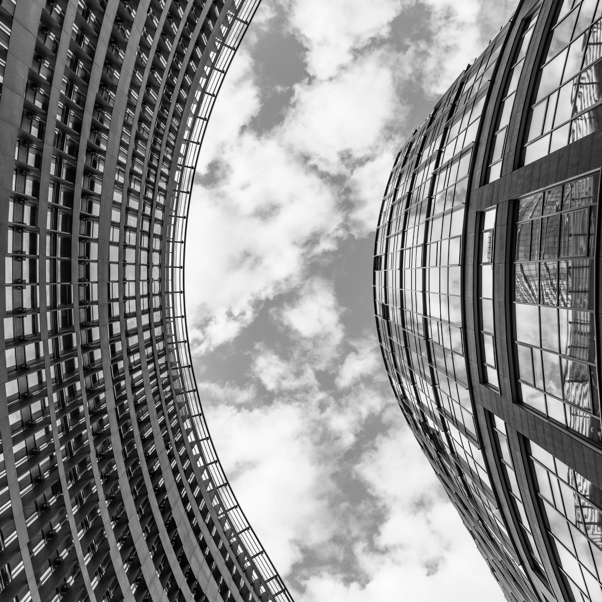 Monochromatyczne ujęcie z dołu na dwa zakrzywione budynki biurowe, między którymi widoczne są chmury. Fasady budynków z licznymi oknami tworzą geometryczny wzór.