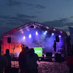 Scena plenerowa z namiotem oświetlonym na czerwono i niebiesko, DJ za konsoletą z logo 'Framex', publiczność na piaszczystej plaży. Wieczorny event.