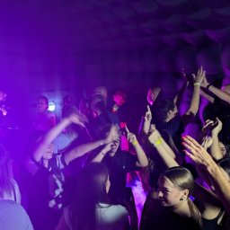 N-Event - Roztańczony tłum w fioletowym oświetleniu, widoczni DJ-e w tle, podniesione ręce i dynamiczne pozy, impreza w klubie z charakterystyczną ścianą.