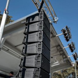 N-Event - Zestaw profesjonalnych głośników liniowych zawieszonych na metalowej konstrukcji kratownicowej, oświetlenie sceniczne w tle, błękitne niebo.