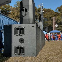 N-Event - Rząd czarnych kolumn głośnikowych na trawie, z widocznym fragmentem sceny i publicznością w tle, oświetlonych naturalnym światłem słonecznym.