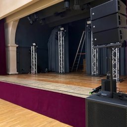 N-Event - Scena z nagłośnieniem i oświetleniem na statywach, drabina w tle. Parkietowa podłoga i bordowa dekoracja sceniczna. Przygotowania do wydarzenia.