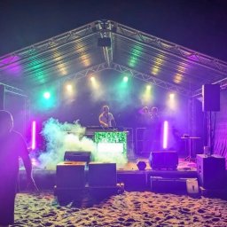 N-Event - Scena plenerowa z DJ-em i oświetleniem w kolorach zieleni i fioletu, z efektem dymu. Widoczni ludzie na scenie i przed nią, piasek na podłożu.