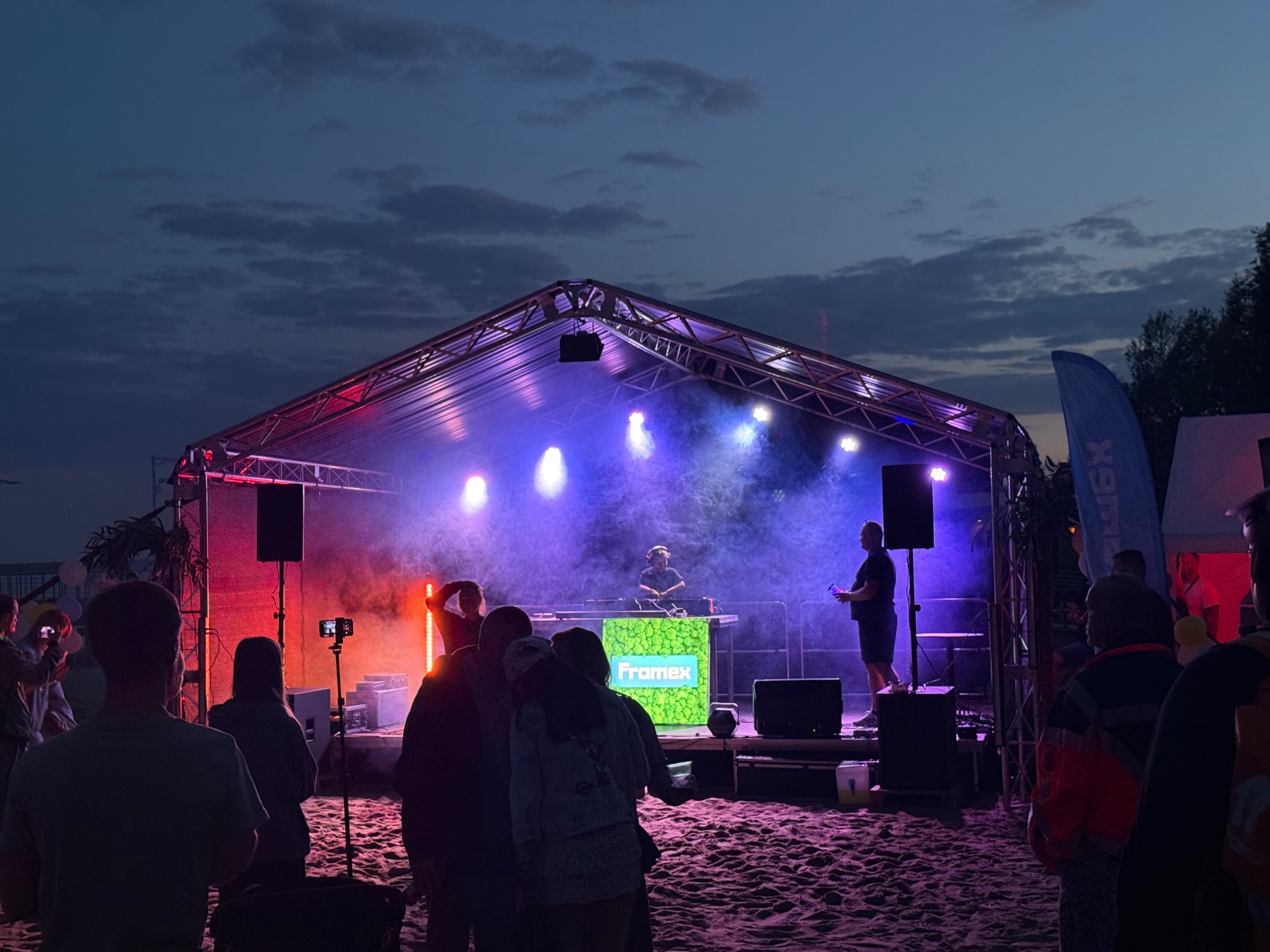 Scena plenerowa z namiotem oświetlonym na czerwono i niebiesko, DJ za konsoletą z logo 'Framex', publiczność na piaszczystej plaży. Wieczorny event.