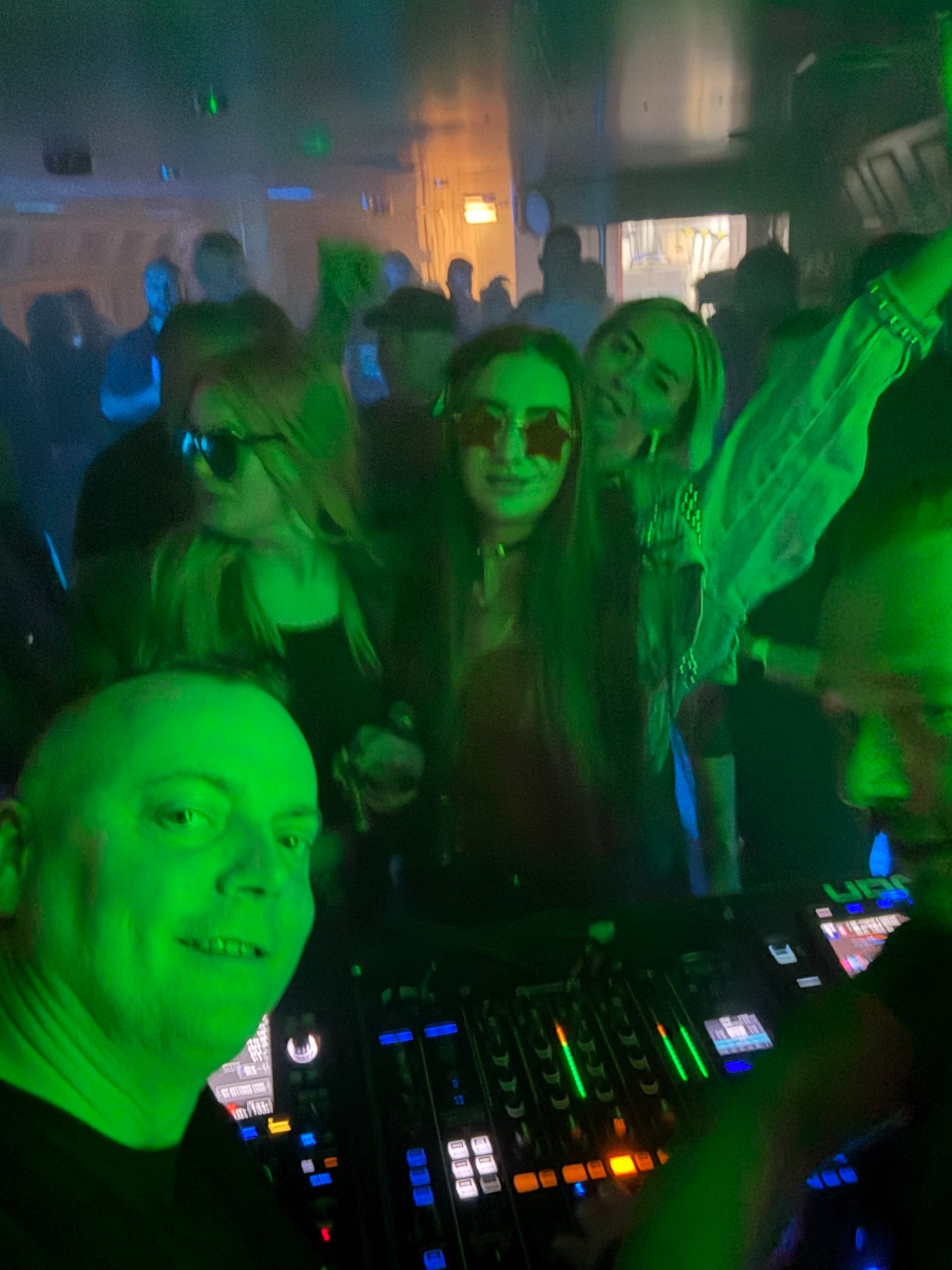 DJ z uśmiechem na twarzy, mikser i rozbawiony tłum w zielonym oświetleniu klubu. Dziewczyna w okularach w kształcie gwiazd.