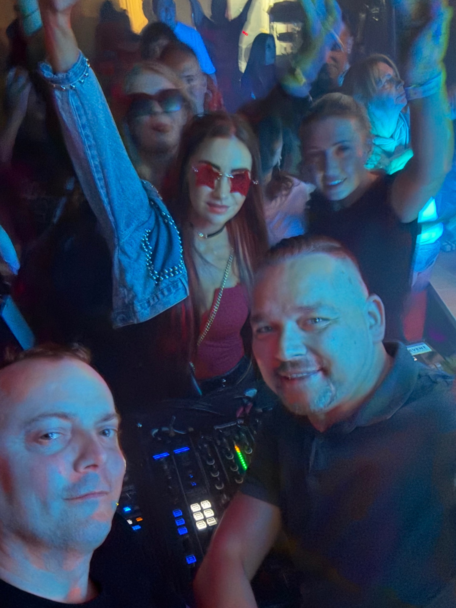 DJ selfie z imprezy: uśmiechnięty DJ z mikserem na tle rozbawionego tłumu w klubie, oświetlenie dyskotekowe, wesoła atmosfera.