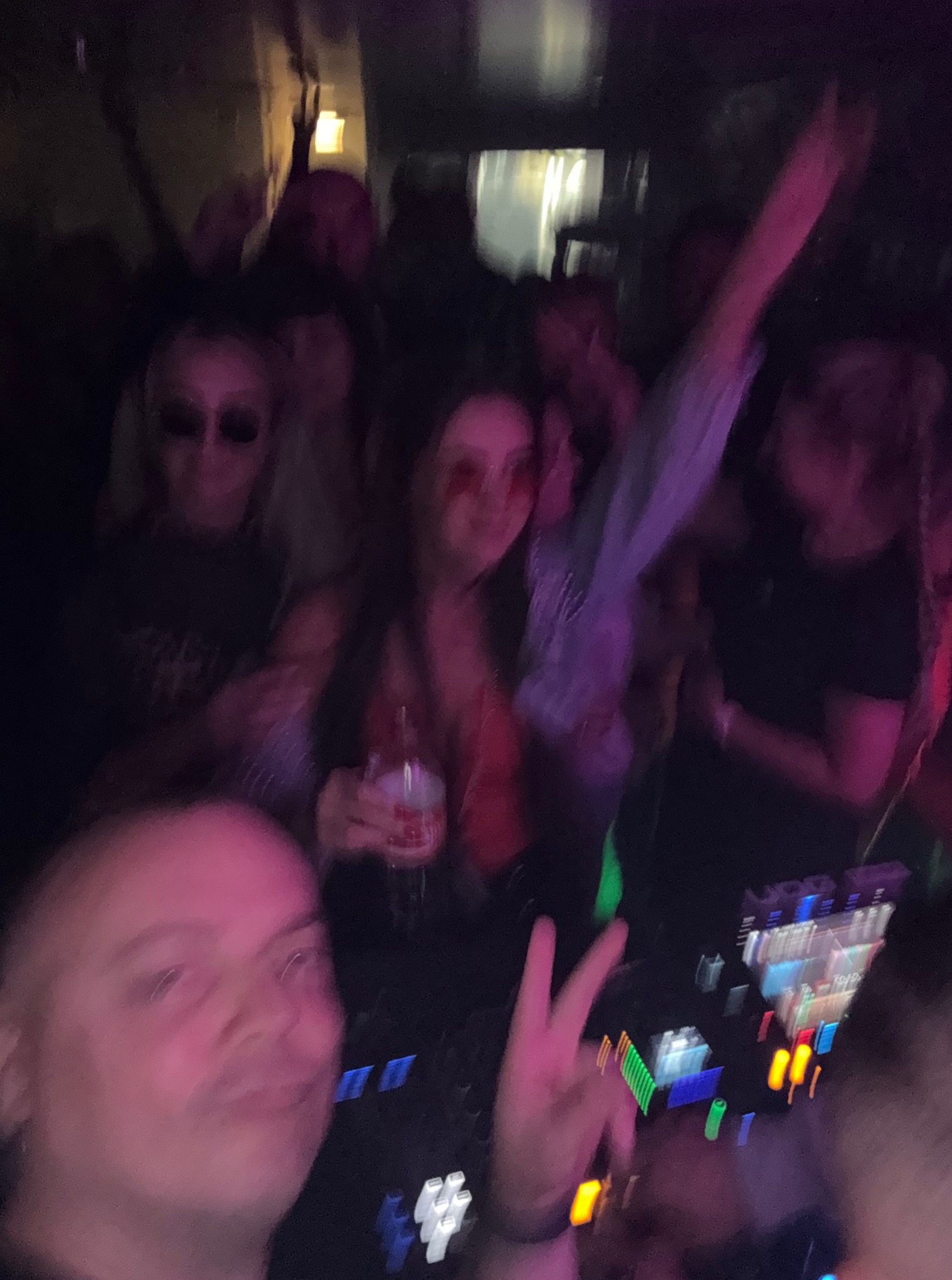 Rozmazane ujęcie z imprezy z DJ-em, rozbawionym tłumem i oświetleniem dyskotekowym. Osoba robi znak V palcami.