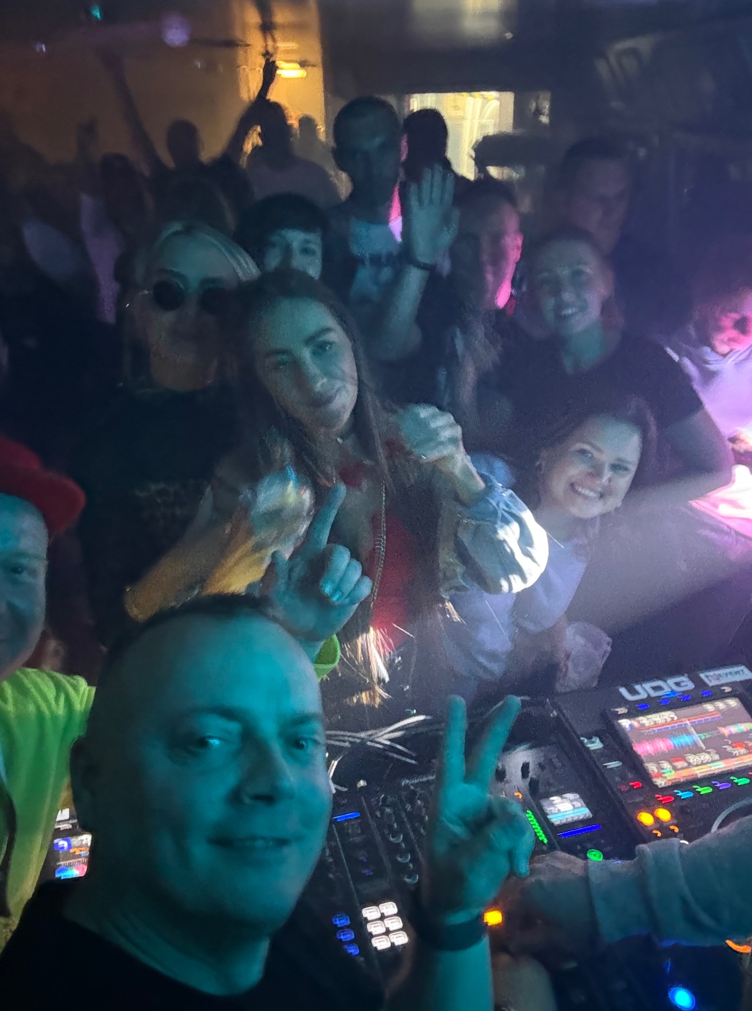 DJ robi selfie z rozbawionym tłumem w klubie. W tle parkiet i oświetlenie dyskotekowe. DJ pokazuje znak zwycięstwa, obok mikser UDG.