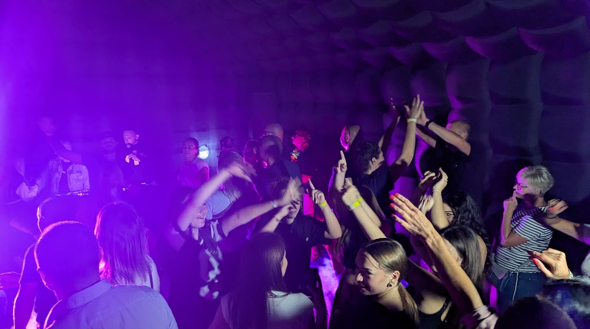 Roztańczony tłum w fioletowym oświetleniu, widoczni DJ-e w tle, podniesione ręce i dynamiczne pozy, impreza w klubie z charakterystyczną ścianą.