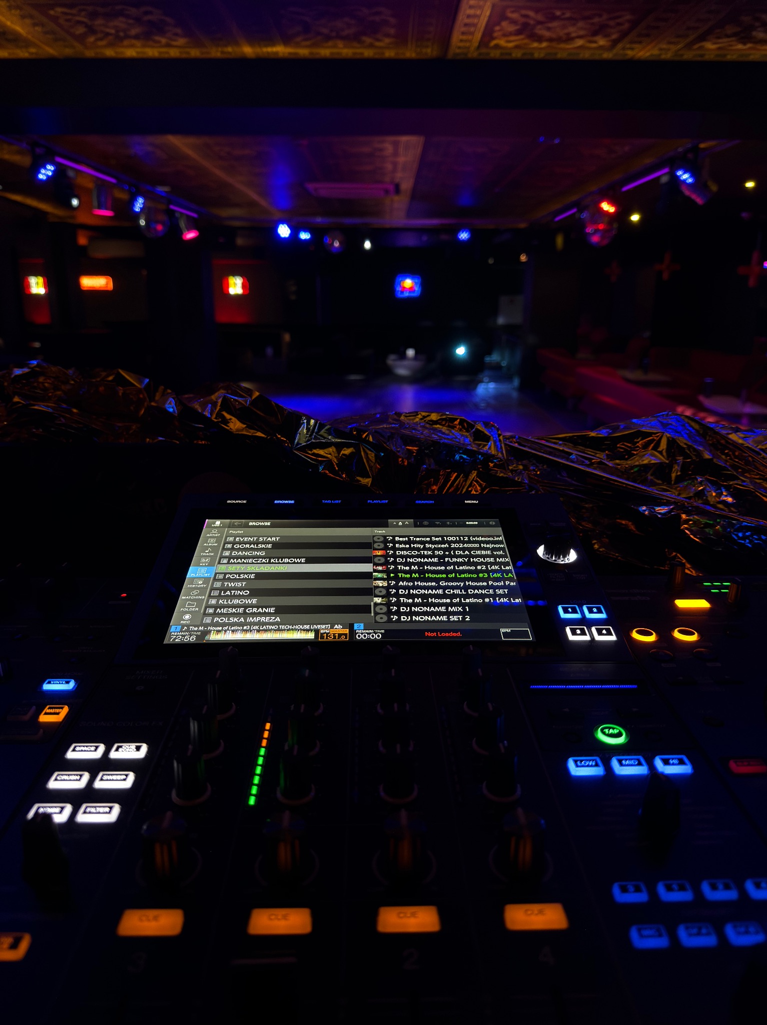 Konsoleta DJ-ska z ekranem dotykowym, podświetlona w klubowym otoczeniu. Widoczne efekty świetlne i lista utworów na ekranie.