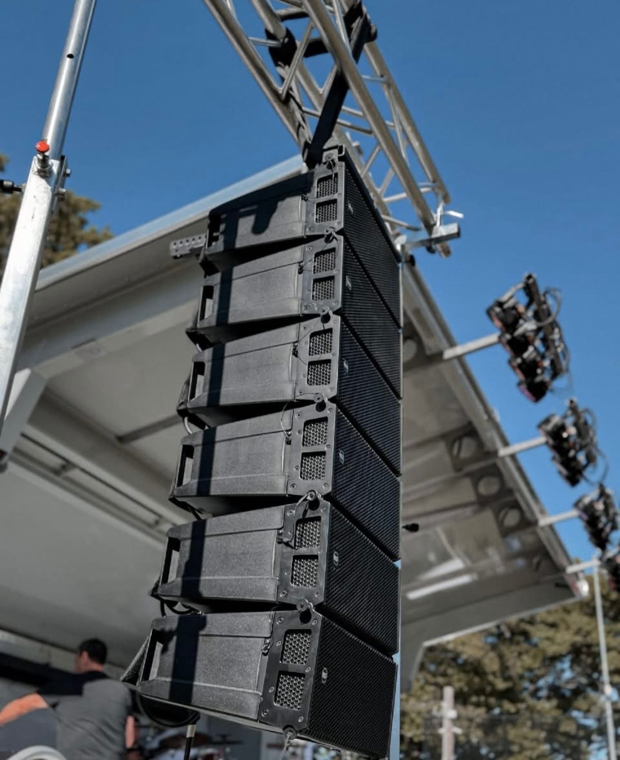 Zestaw profesjonalnych głośników liniowych zawieszonych na metalowej konstrukcji kratownicowej, oświetlenie sceniczne w tle, błękitne niebo.