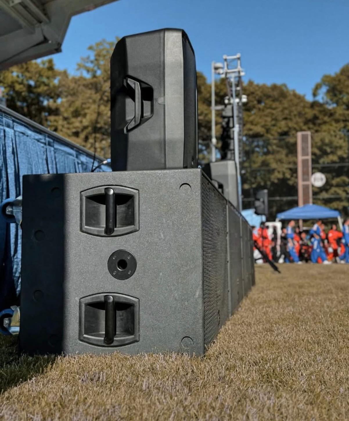Rząd czarnych kolumn głośnikowych na trawie, z widocznym fragmentem sceny i publicznością w tle, oświetlonych naturalnym światłem słonecznym.