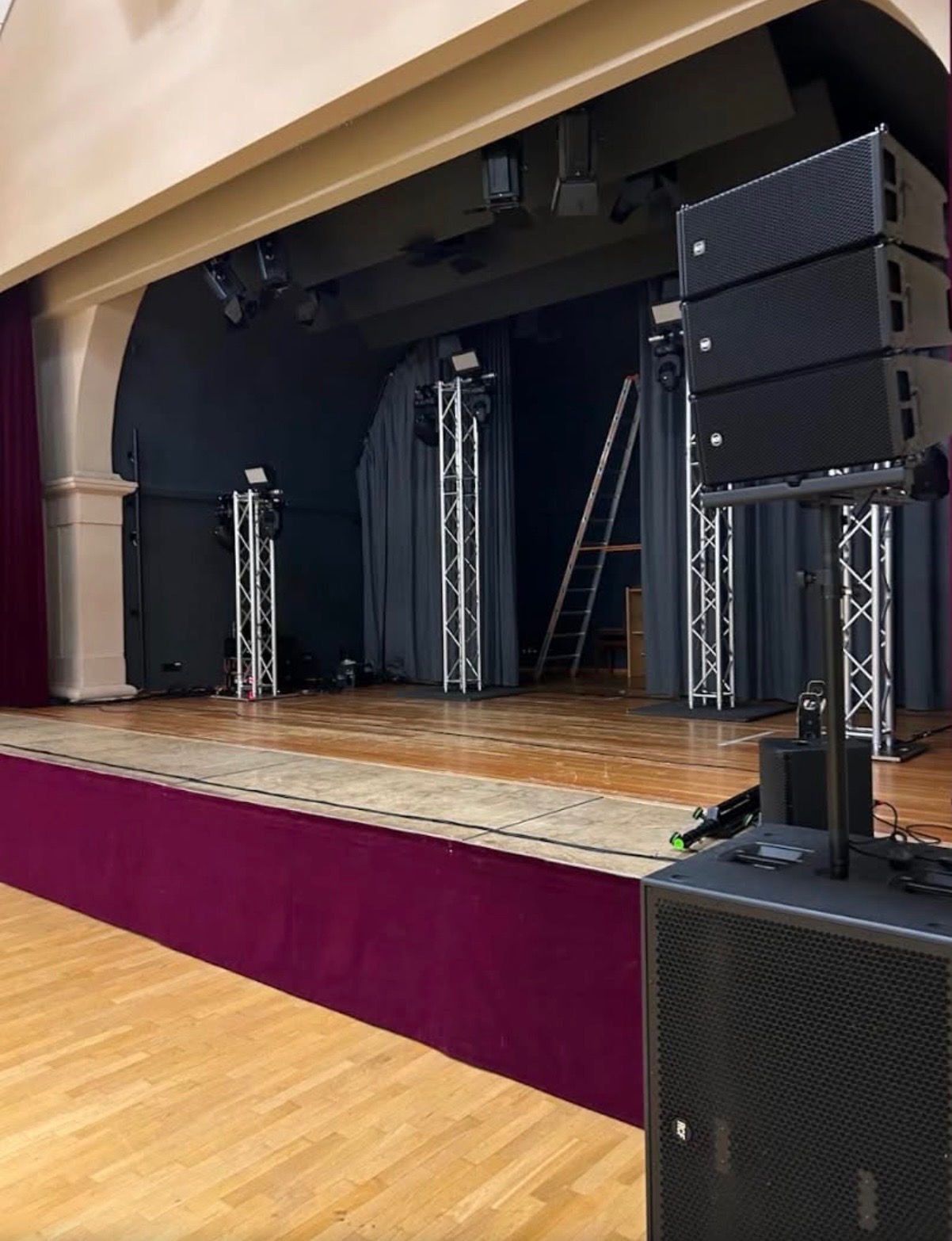 Scena z nagłośnieniem i oświetleniem na statywach, drabina w tle. Parkietowa podłoga i bordowa dekoracja sceniczna. Przygotowania do wydarzenia.