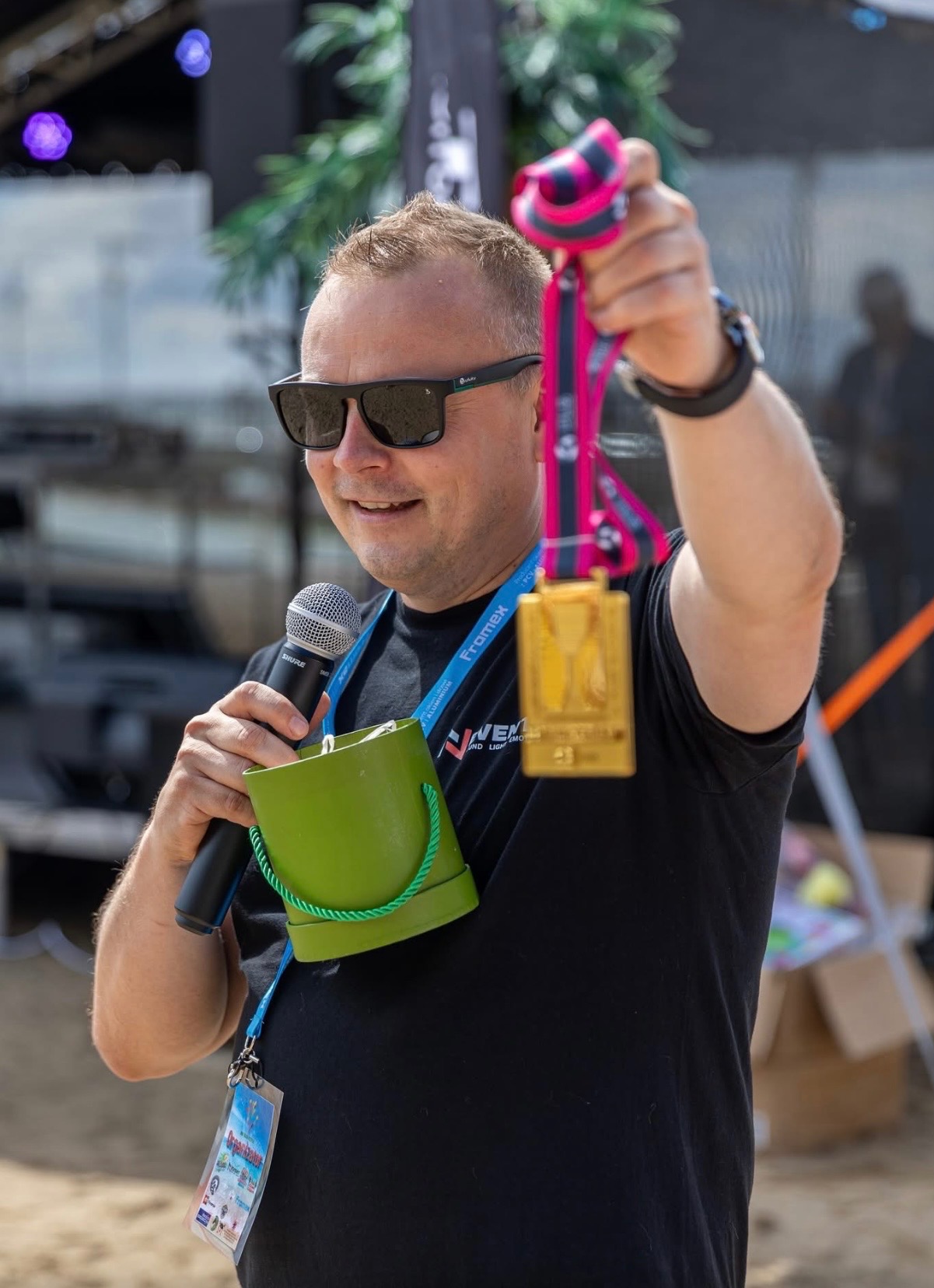 Uśmiechnięty mężczyzna w okularach, z mikrofonem i złotym medalem na tle sceny. Nietypowy zielony pojemnik na mikrofon.